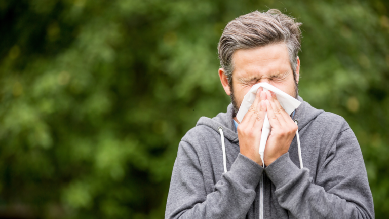 Man in a gray hoodie sneezes into a tissue outdoors, eyes closed in motion blur of greenery behind him.