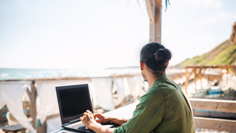 Person typing on a laptop at an outdoor beachside cafe with the ocean in the background