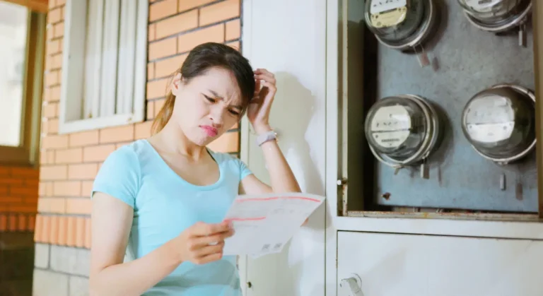 Young woman stands outside a brick building, squinting at a document she’s reading near a utility meter panel on the wall