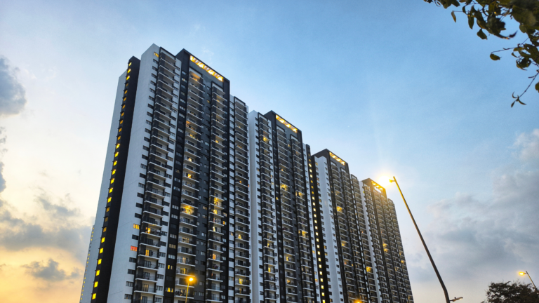 Tall modern apartment complex at dusk with lit windows and streetlights along the foreground.