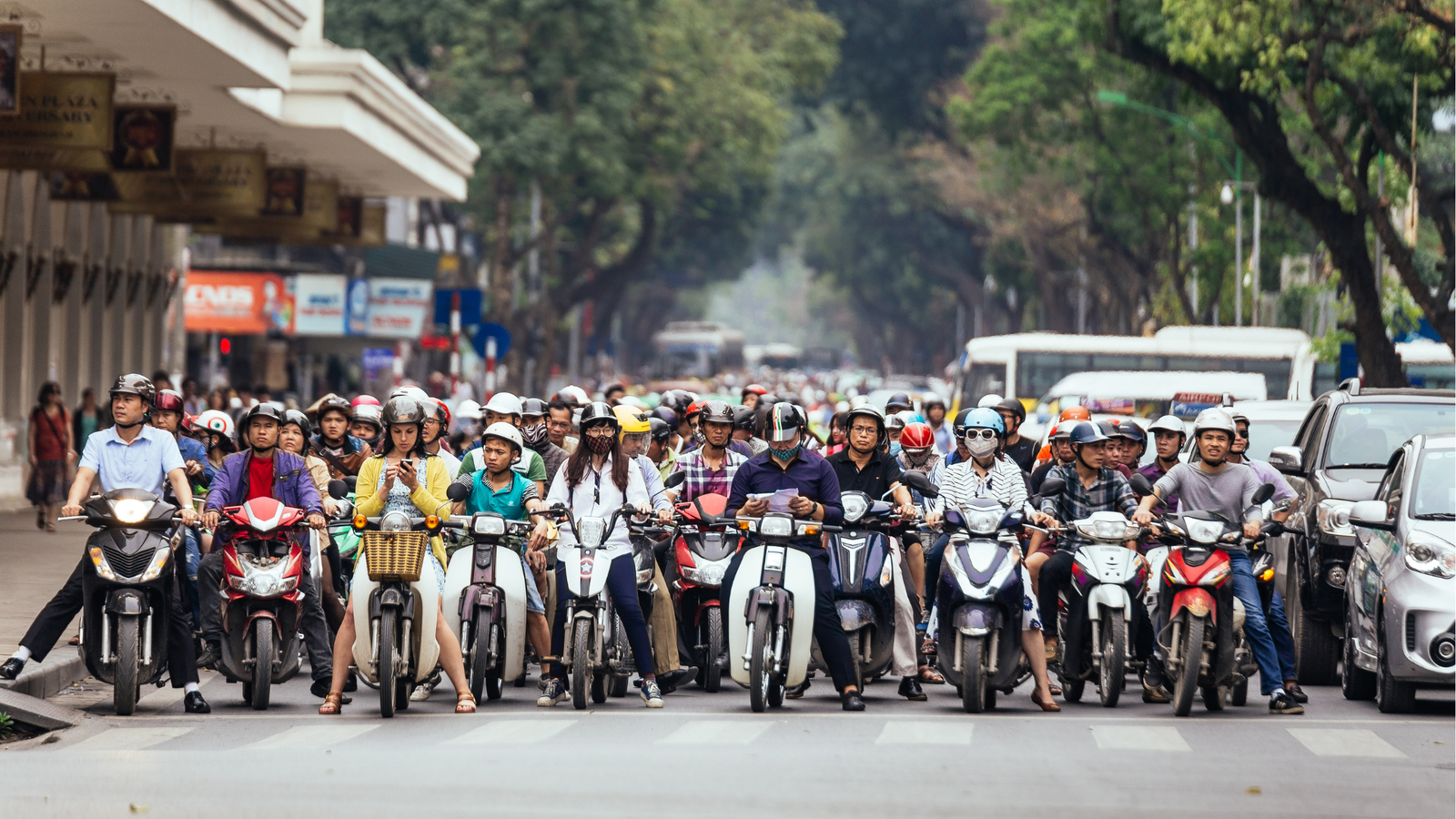 Crowded city street filled with many motorbike riders wearing helmets; cars and buses in the background.