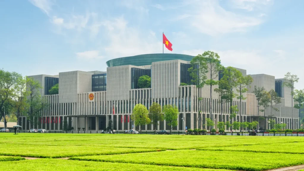 Modern government building with a Vietnam flag on a tall pole, surrounded by green trees and a wide manicured lawn in the foreground
