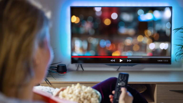 Person reclining on a sofa with popcorn, watching a blurred cityscape on a flat-screen TV, holding a remote.